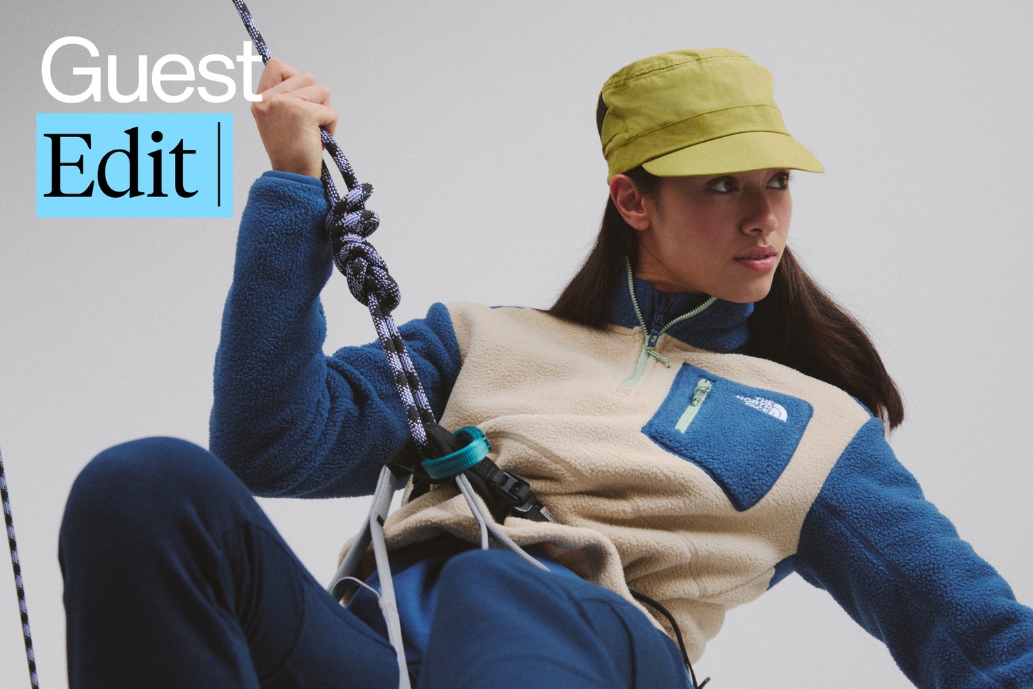 A person wearing a blue and beige fleece jacket and green cap grips a climbing rope, focused on their ascent against a gray background.