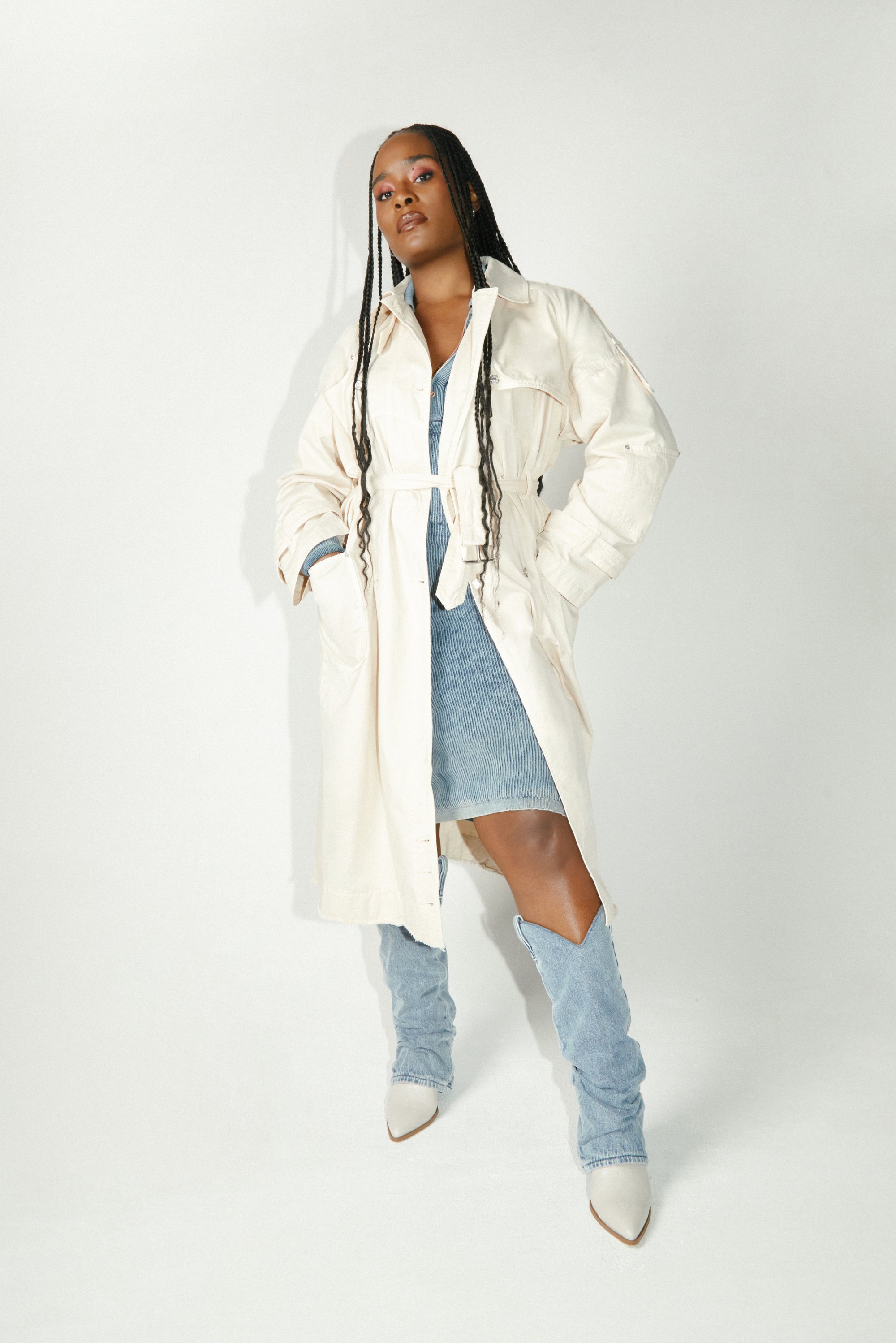 Una donna con lunghi capelli intrecciati posa indossando un leggero trench coat sopra un vestito di jeans e stivali blu morbidi, contro uno sfondo bianco.