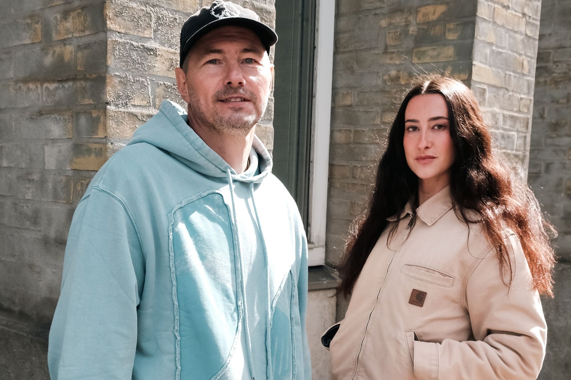 Två individer står utomhus mot en tegelvägg. Mannen har på sig en ljusblå huvtröja. Kvinnan har en beige jacka med ett märke.