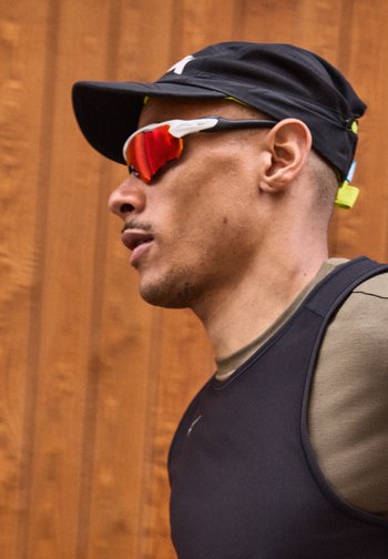 Άνδρας που φοράει μαύρο καπέλο, κόκκινα αθλητικά γυαλιά ηλίου με ανακλαστικούς φακούς και μαύρο αμάνικο αθλητικό μπλουζάκι, στέκεται μπροστά σε ξύλινο φόντο.