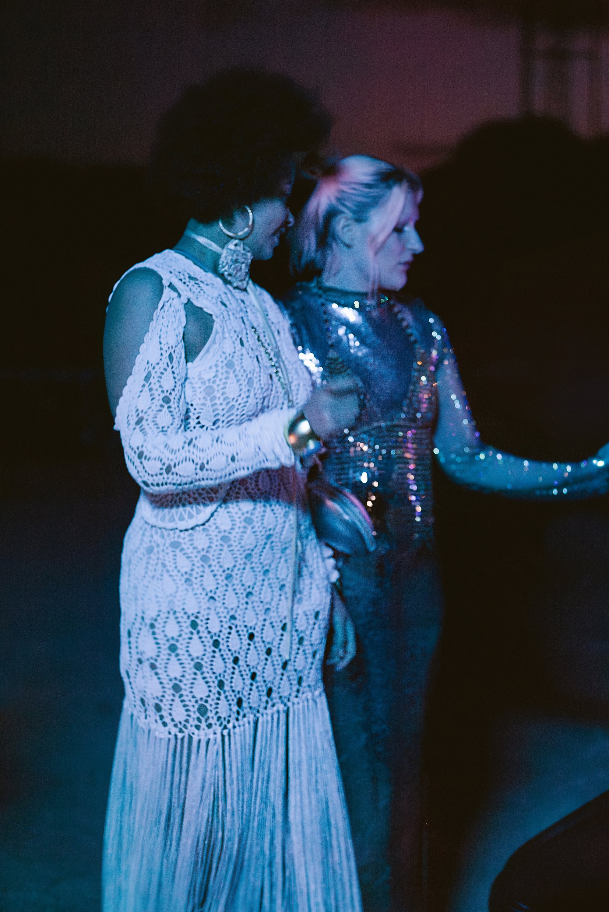 Two women engage in conversation; one in a white, lace dress, the other in a sparkling silver outfit, under dim, colorful lighting.