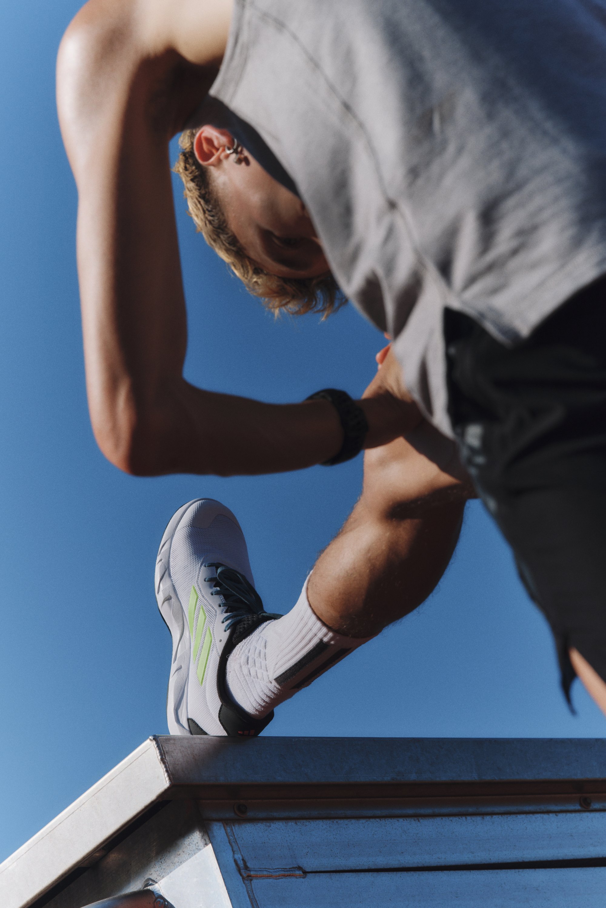 Sportieve schoen in grijs met groene accenten, met een gestructureerd oppervlak en zwarte veters. Geplaatst op een metallic oppervlak onder een blauwe lucht.