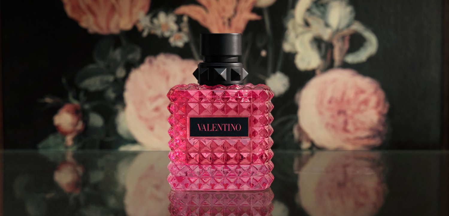 Pink Valentino perfume bottle with textured diamond pattern and black cap on a reflective surface with floral background.