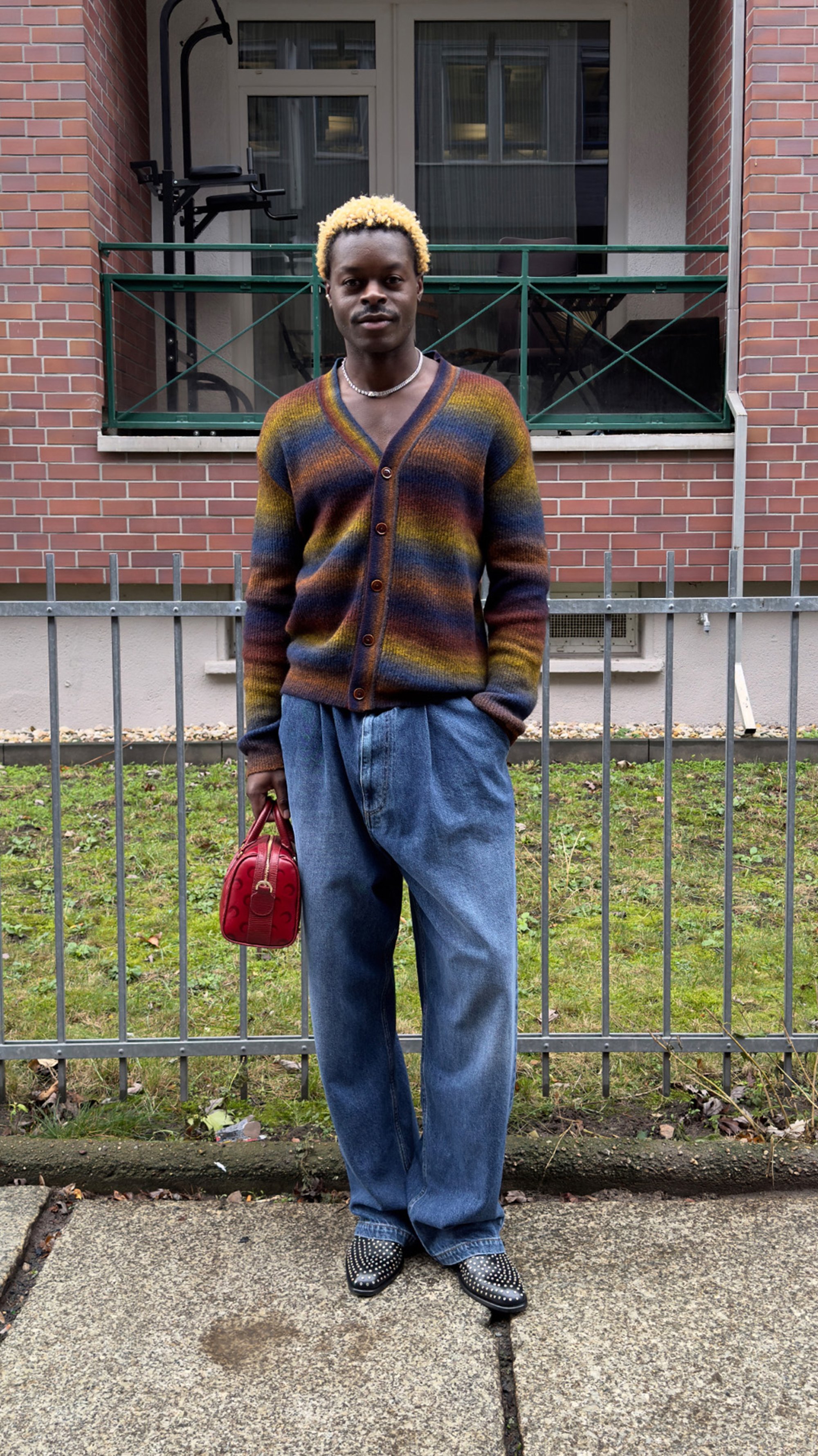 Una persona con cabello rubio rizado lleva un cardigan a rayas colorido y pantalones azules holgados, sosteniendo un pequeño bolso rojo, de pie junto a una cerca.