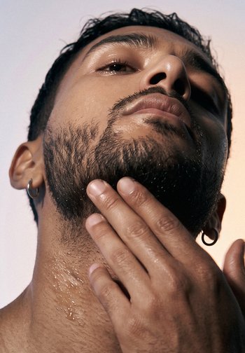 Man with a beard and earring touching his wet neck and chin, looking upward against a plain background.