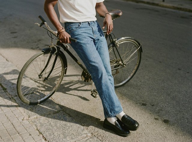 Mężczyzna ubrany w białą koszulkę i niebieskie jeansy opiera się o czarny vintage rower na słonecznym chodniku.