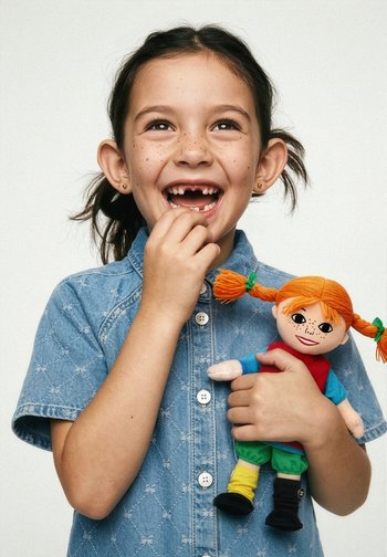 Nasmejana mlada deklica z manjkajočimi sprednjimi zobmi, oblečena v denim srajco, drži barvito lutko z oranžnimi kitkami na enobarvnem ozadju.