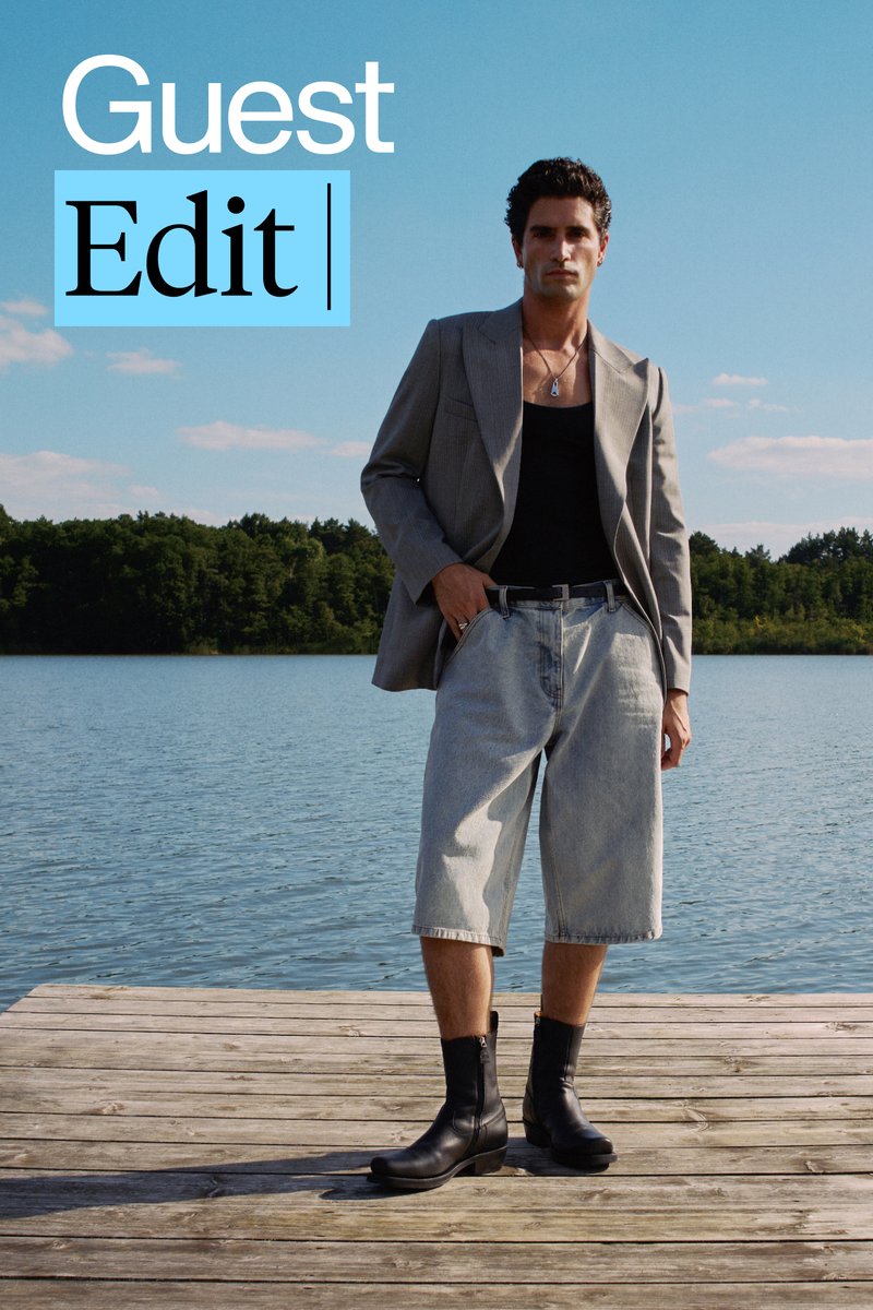 Een man staat op een houten steiger bij een meer, gekleed in een grijze blazer, een zwart hemdtop, oversized denim shorts en zwarte enkellaarzen.