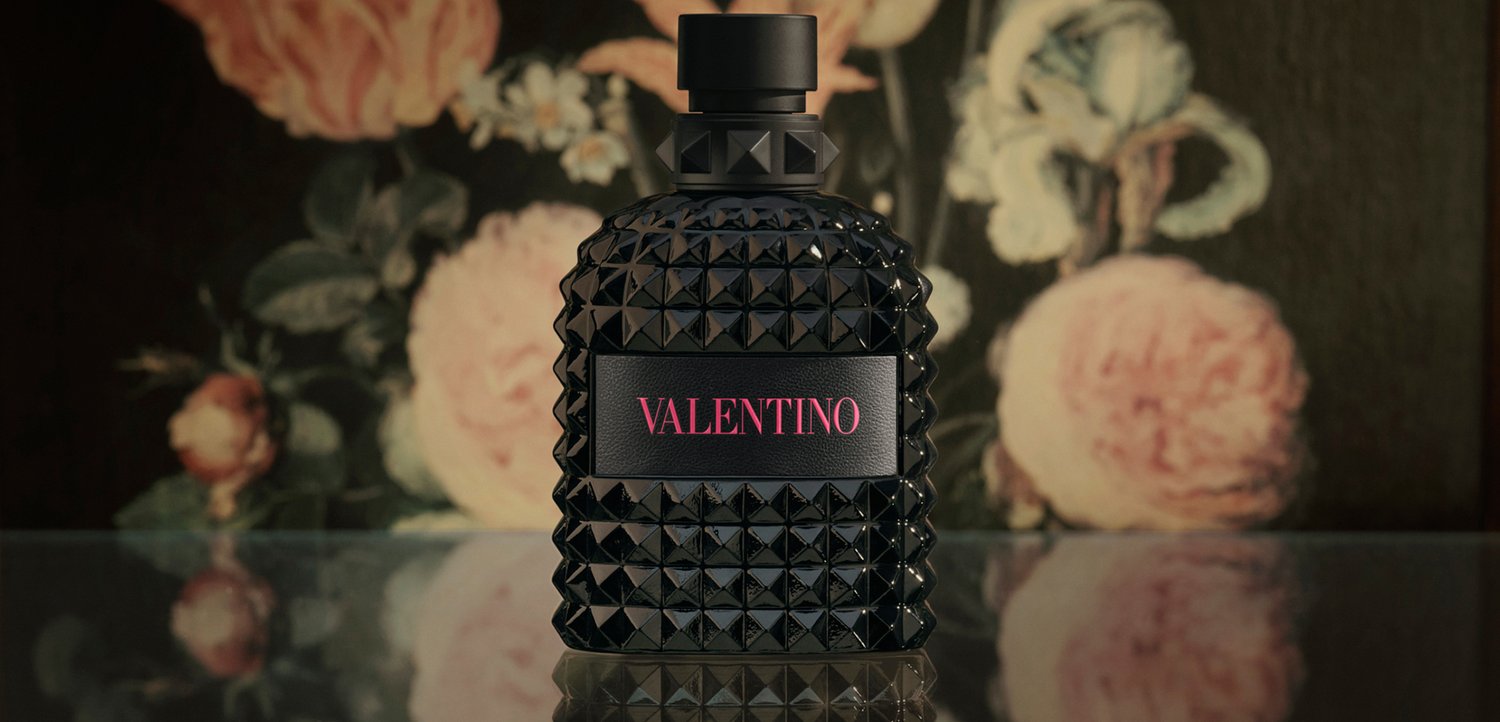 Black studded Valentino perfume bottle with pink text on label, placed on reflective surface with blurred floral background.