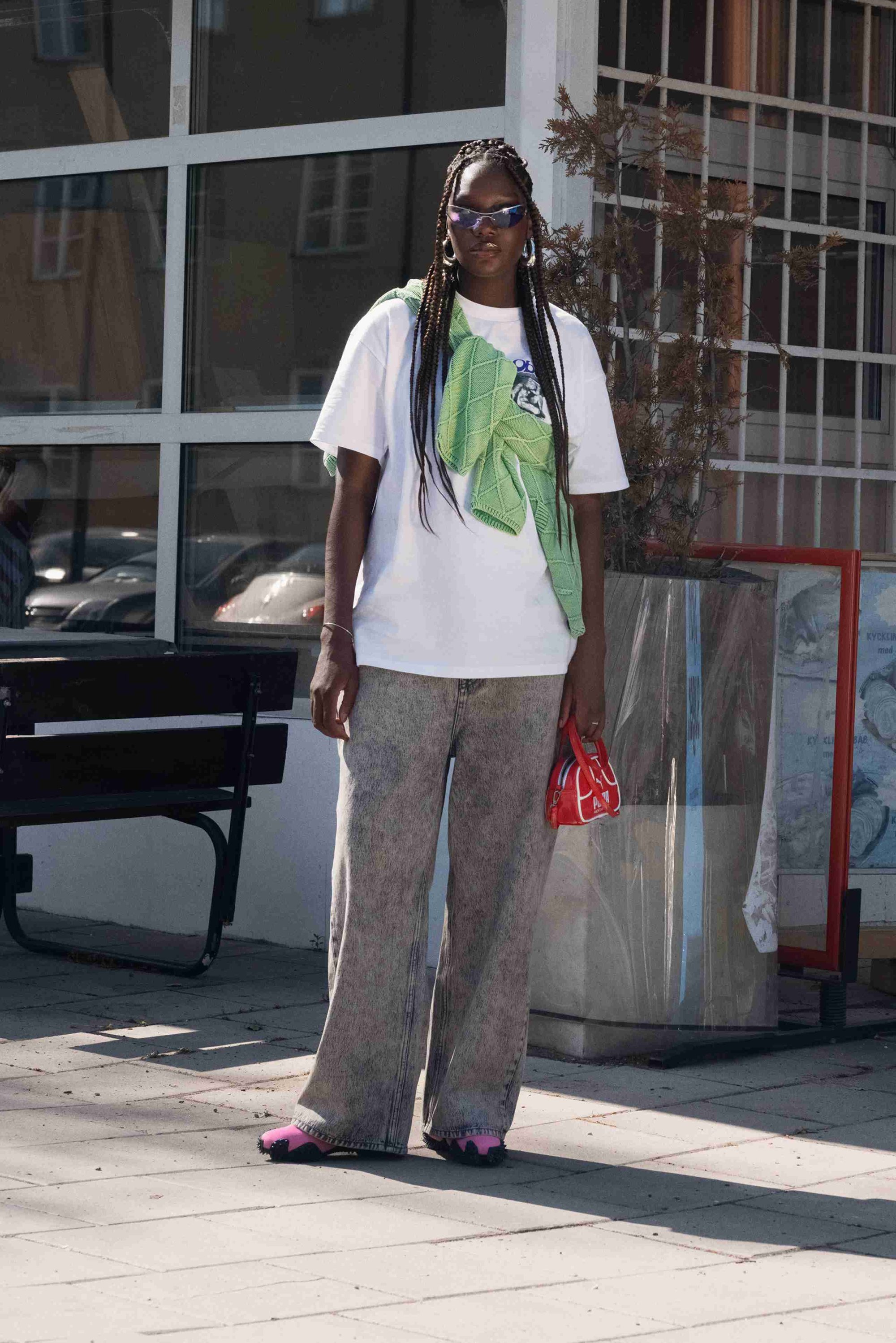 Model wearing a white t-shirt, patterned loose gray jeans, pink footwear, with a green knit shawl draped over shoulders, holding a red handbag.