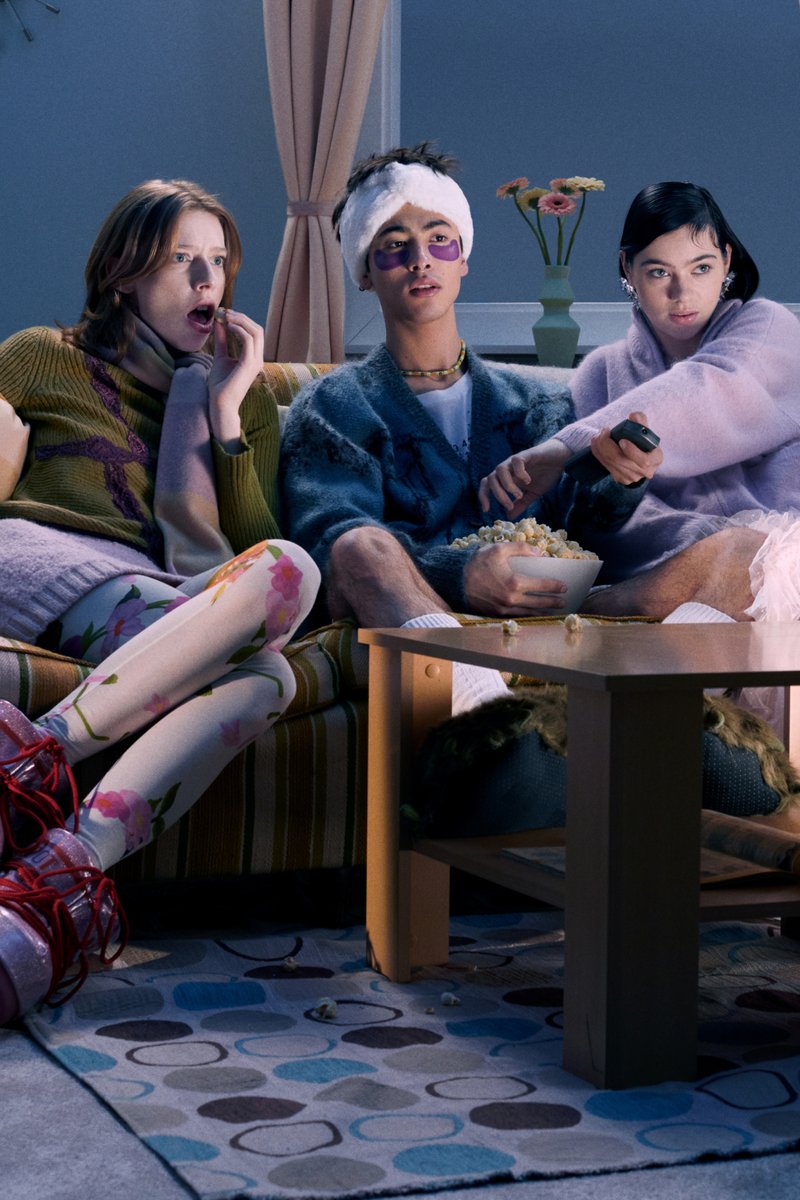 Three friends sit on a striped couch, surprised, watching TV. One holds a remote, another has a bowl of popcorn, colorful outfits highlighted.