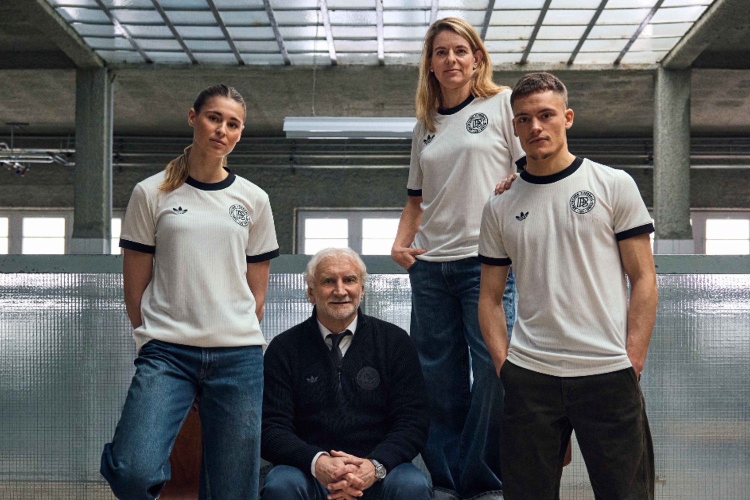 Un gruppo di quattro persone che indossa magliette bianche Adidas con colletto nero, abbinate a jeans e pantaloni scuri. Sfondo industriale.
