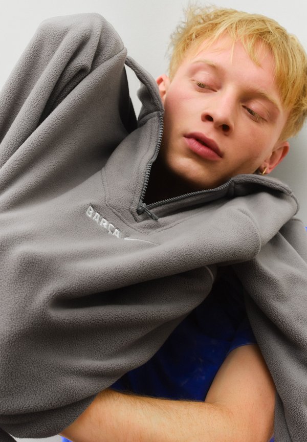 Sivý fleecový pulóver s polovičným zipsom a textúrou látky, ktorý má logo a priliehavý golier, zobrazený na neutrálnom pozadí.