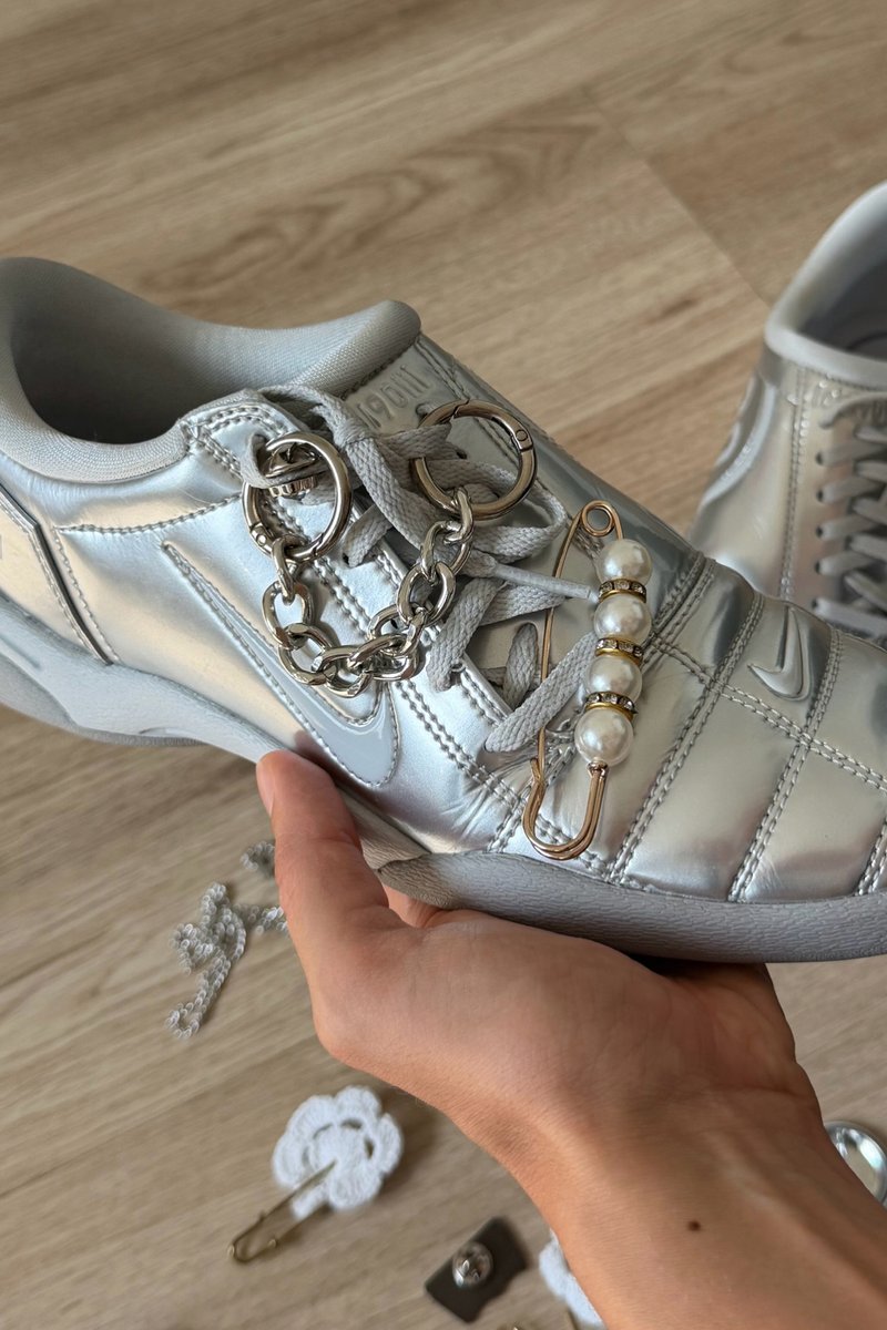 Mano che tiene una sneaker argento con lacci grigi decorata con una catena e una spilla da balia con perline bianche su pavimento in legno.