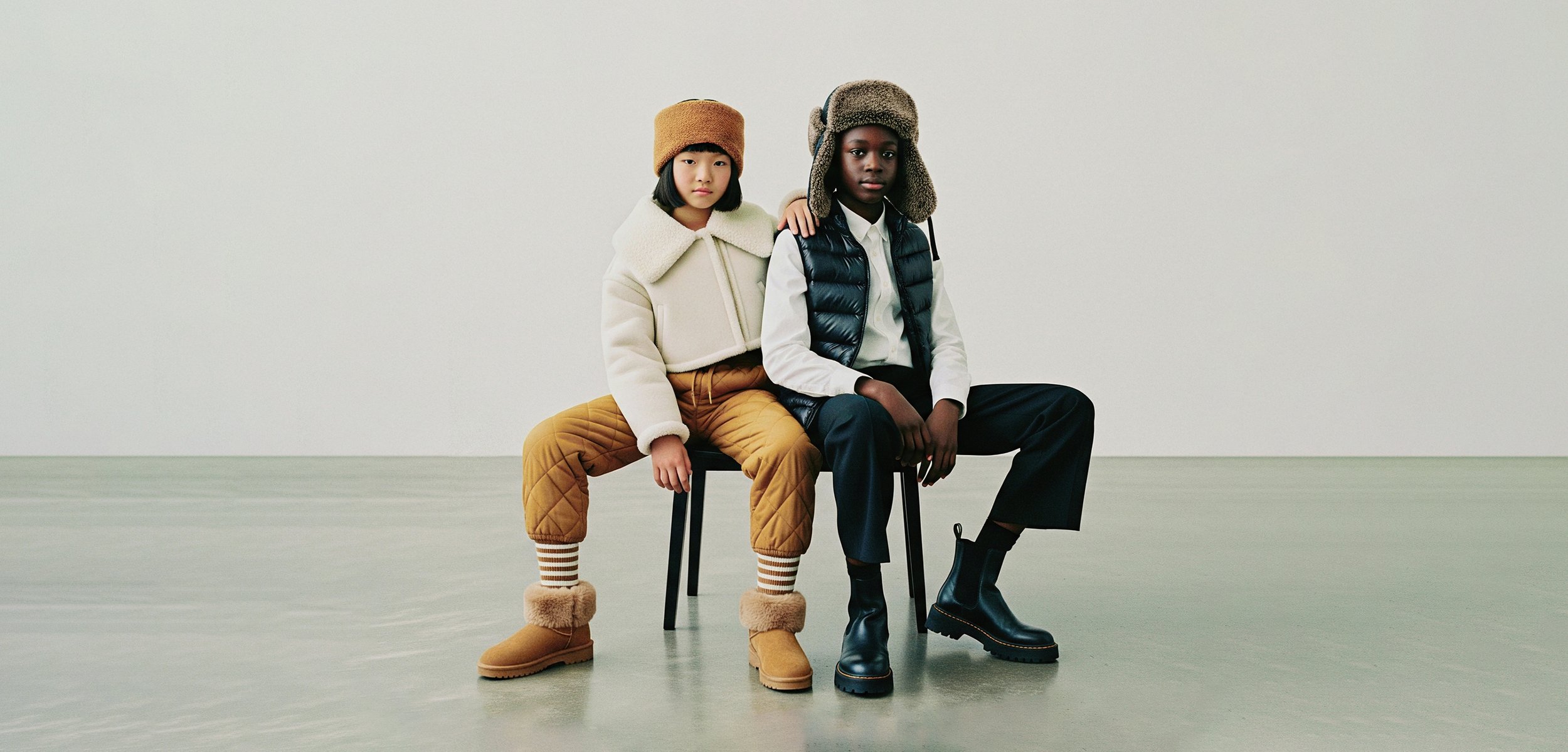 To børn sidder skulder ved skulder på en stol. Den ene har en cremefarvet jakke, en brun hat, quiltede bukser og boots med pelsfor. Den anden har en sort vest, en hvid skjorte og mørke bukser med sorte støvler.