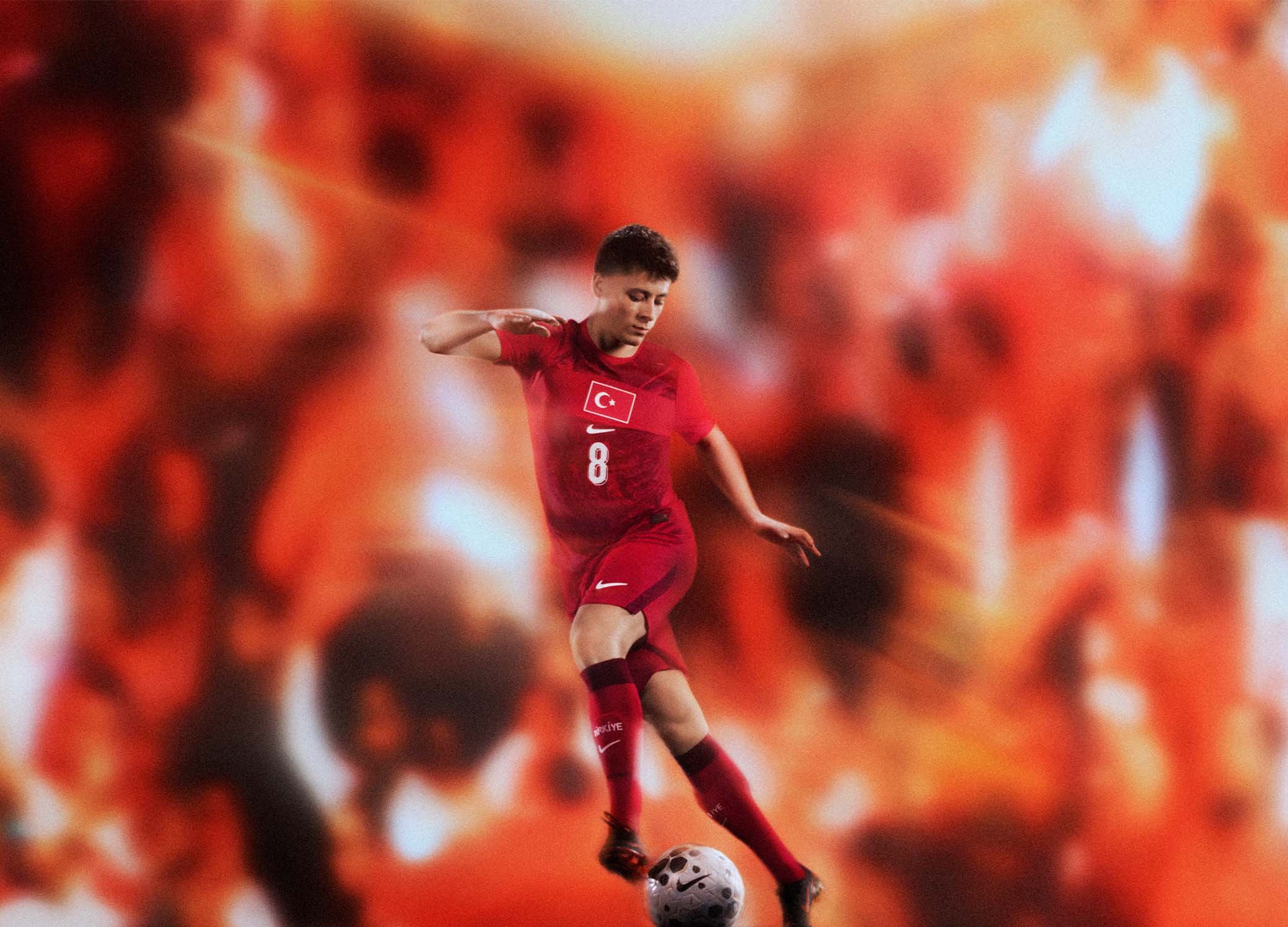 Joueur de football portant l'uniforme rouge de la Turquie, numéro 8, prêt à frapper un ballon noir et blanc sur un fond orange flou.