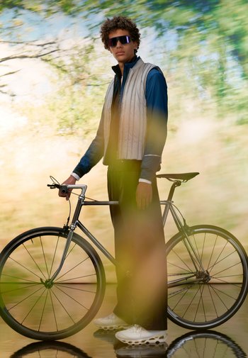 Ein junger Mann in einem stilvollen Outfit steht neben einem Fahrrad, trägt Sonnenbrillen und eine Weste, umgeben von einem verschwommenen natürlichen Hintergrund.