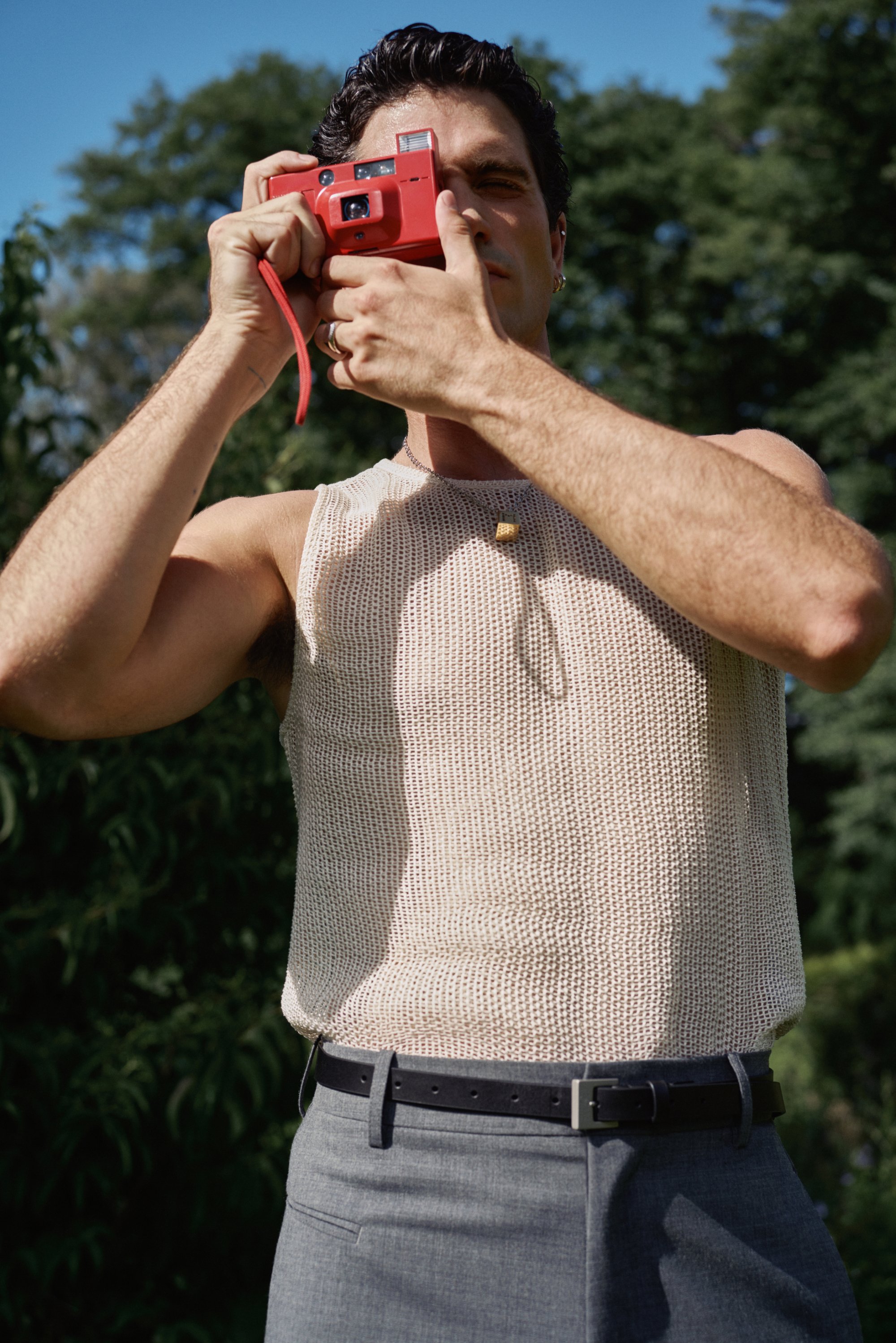 A man in a knitted tank top holds a red camera to his face, surrounded by green foliage under a clear blue sky.
