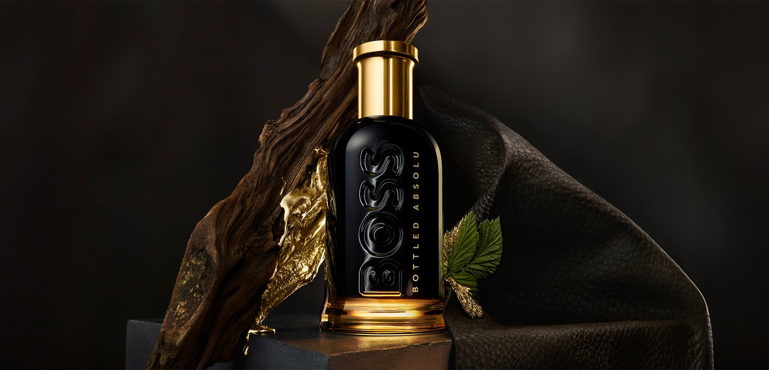Schwarze und goldene BOSS Parfümflasche auf einem Sockel, umgeben von strukturiertem dunklem Stoff, Rinde und einem grünen Blatt.