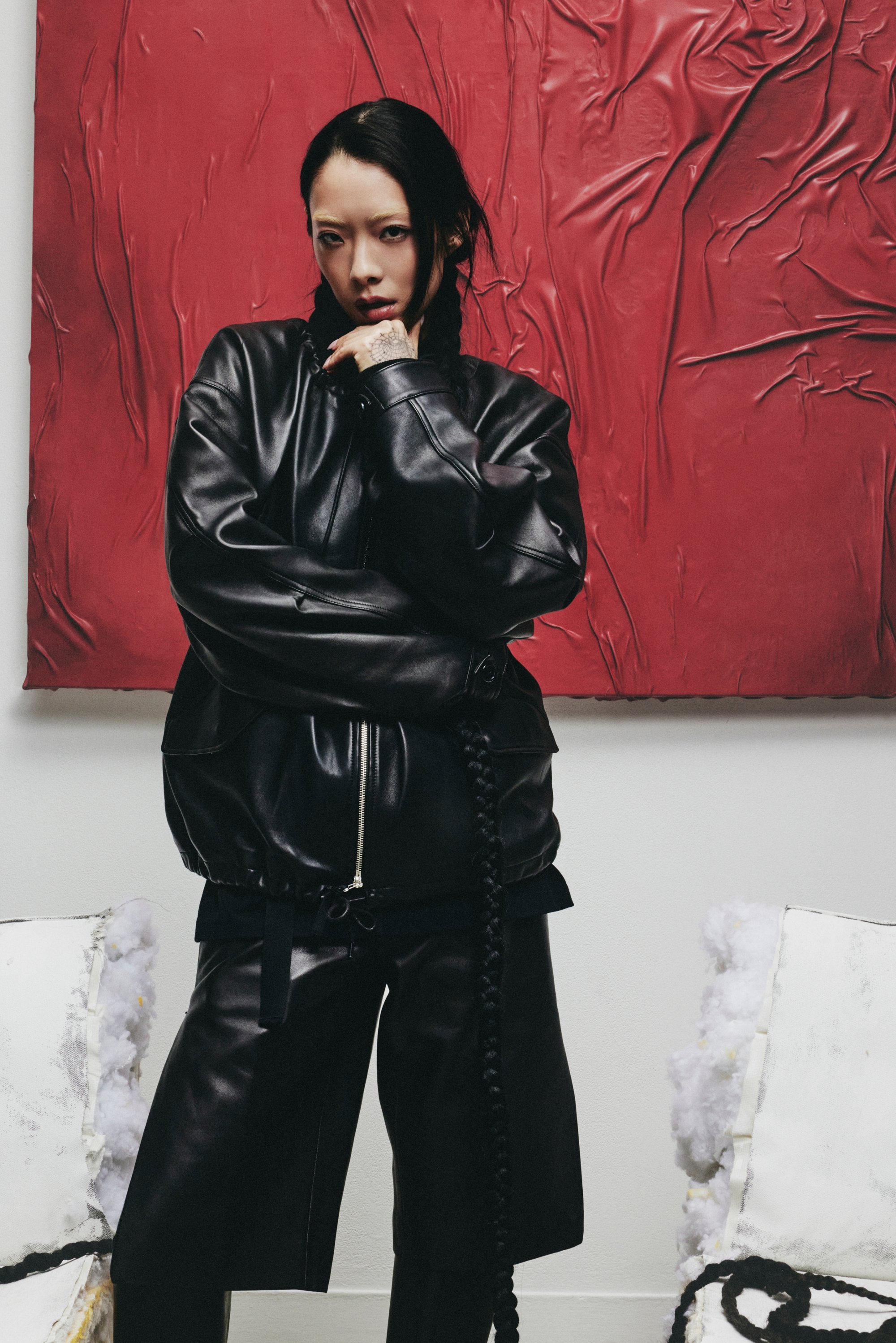 Una persona con una chaqueta de cuero negra y pantalones cortos posa con confianza contra un fondo texturizado rojo, con una silla parcialmente visible a un lado.