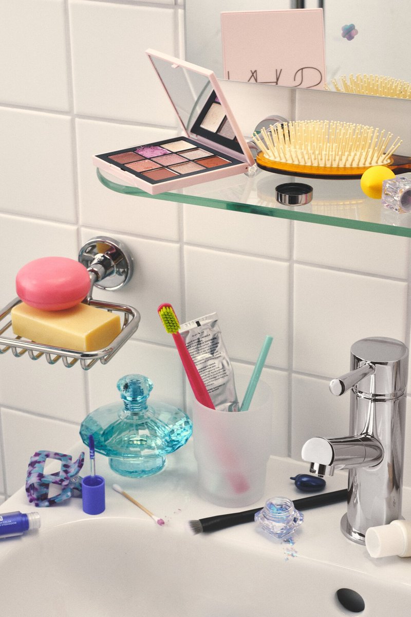 Palette per trucco, spazzola per capelli, spazzolini da denti, dentifricio, saponette, flacone di profumo e articoli cosmetici su un lavabo e una mensola in vetro del bagno.