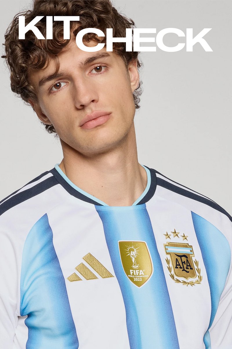 Mladý muž s kudrnatými vlasy, který má na sobě argentinský fotbalový dres s logy FIFA 2022 Mistrovství světa a AFA na hrudi, na jednobarevném pozadí.