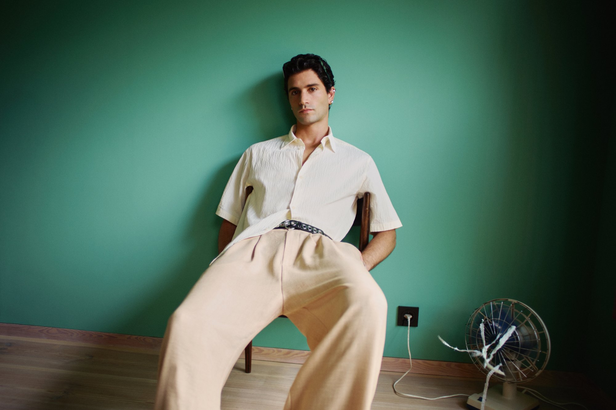 Man sitting casually on a wooden chair against a green wall, wearing a short-sleeved shirt and wide beige pants, with a fan nearby.