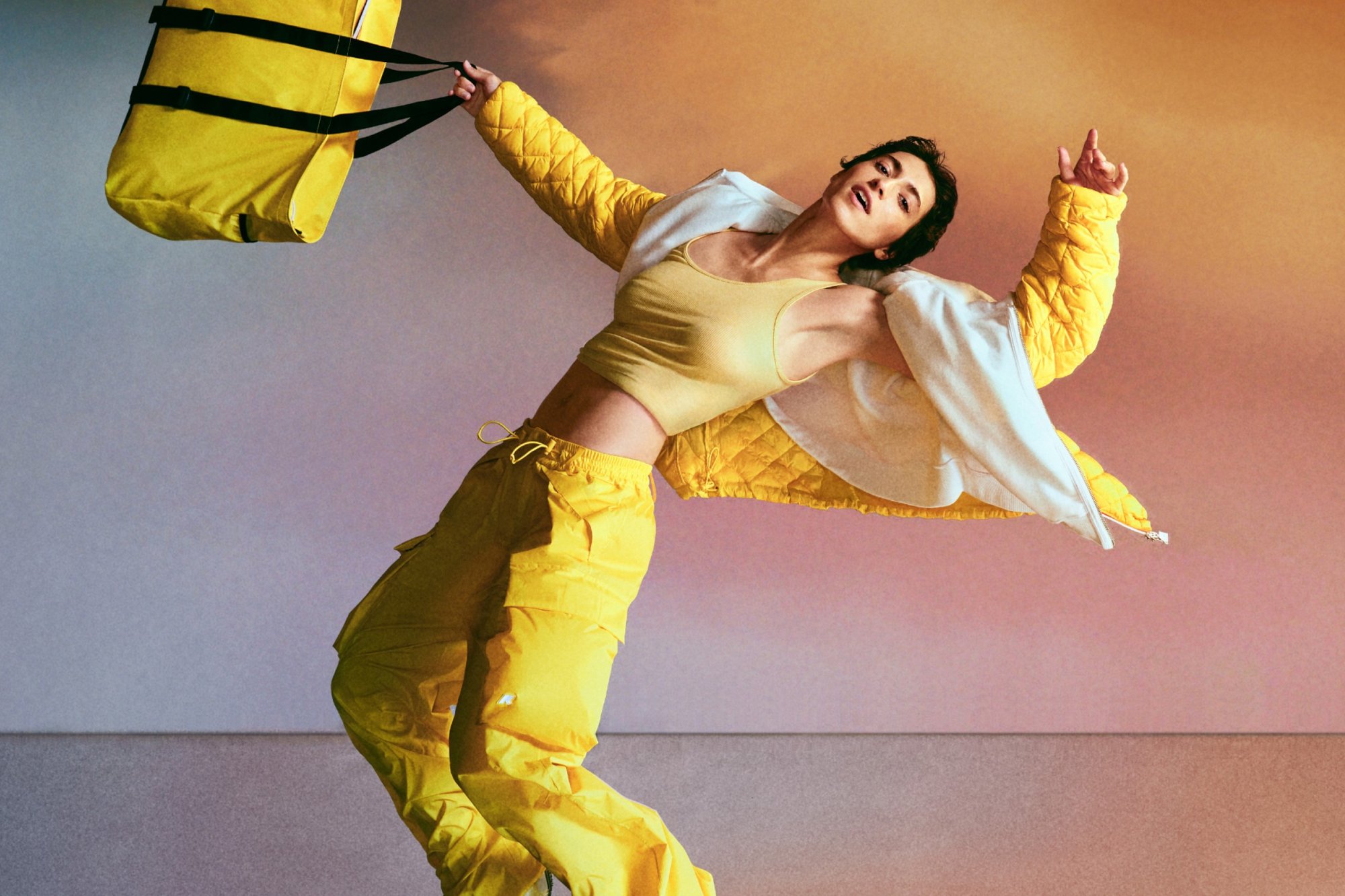 A person in yellow athletic wear and a white jacket dramatically poses, swinging a large yellow bag against a softly colored backdrop.