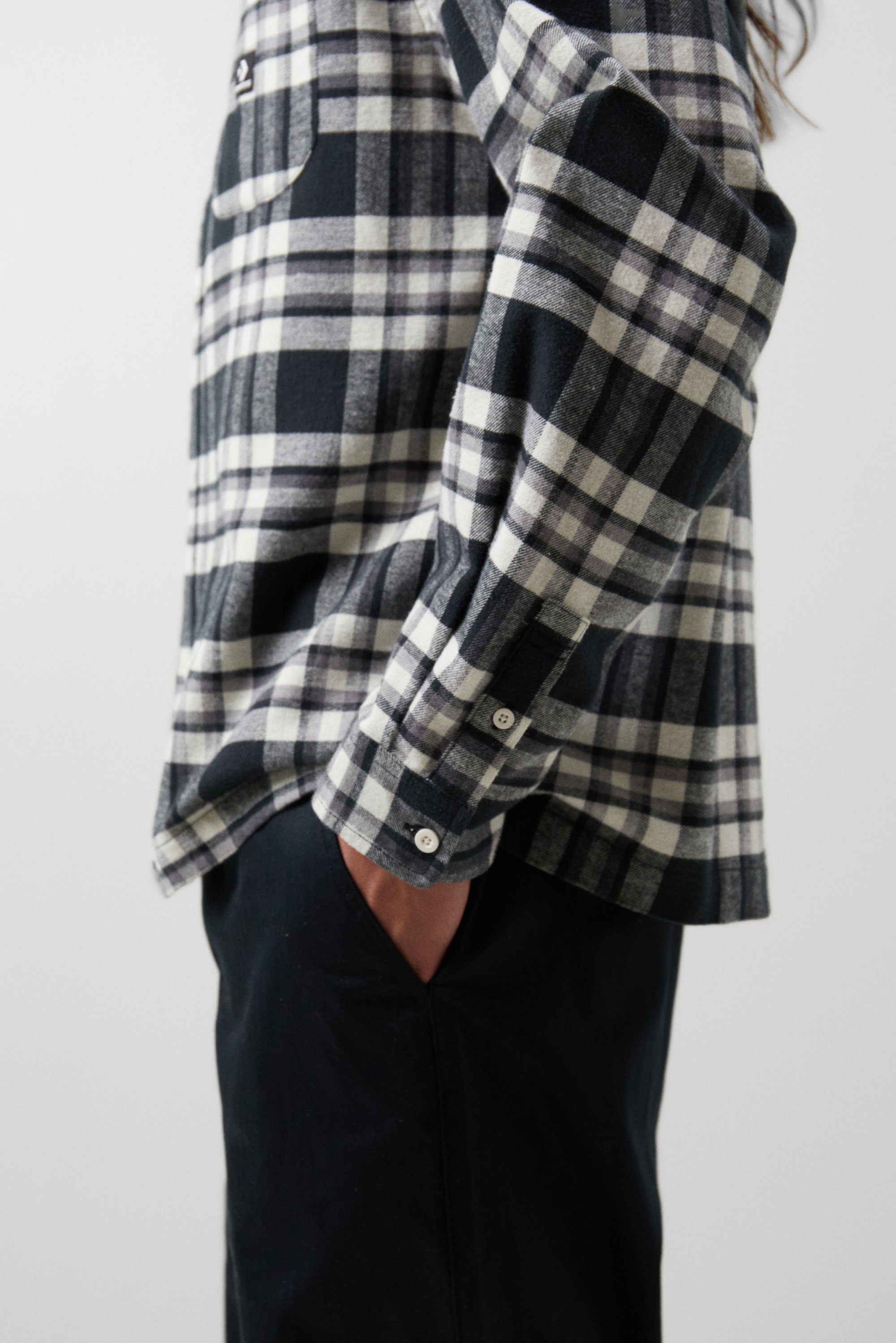 Een persoon in een geblokt zwart en grijs flanellen shirt met opgestroopte mouwen, staand met handen in hun zakken tegen een neutrale achtergrond.