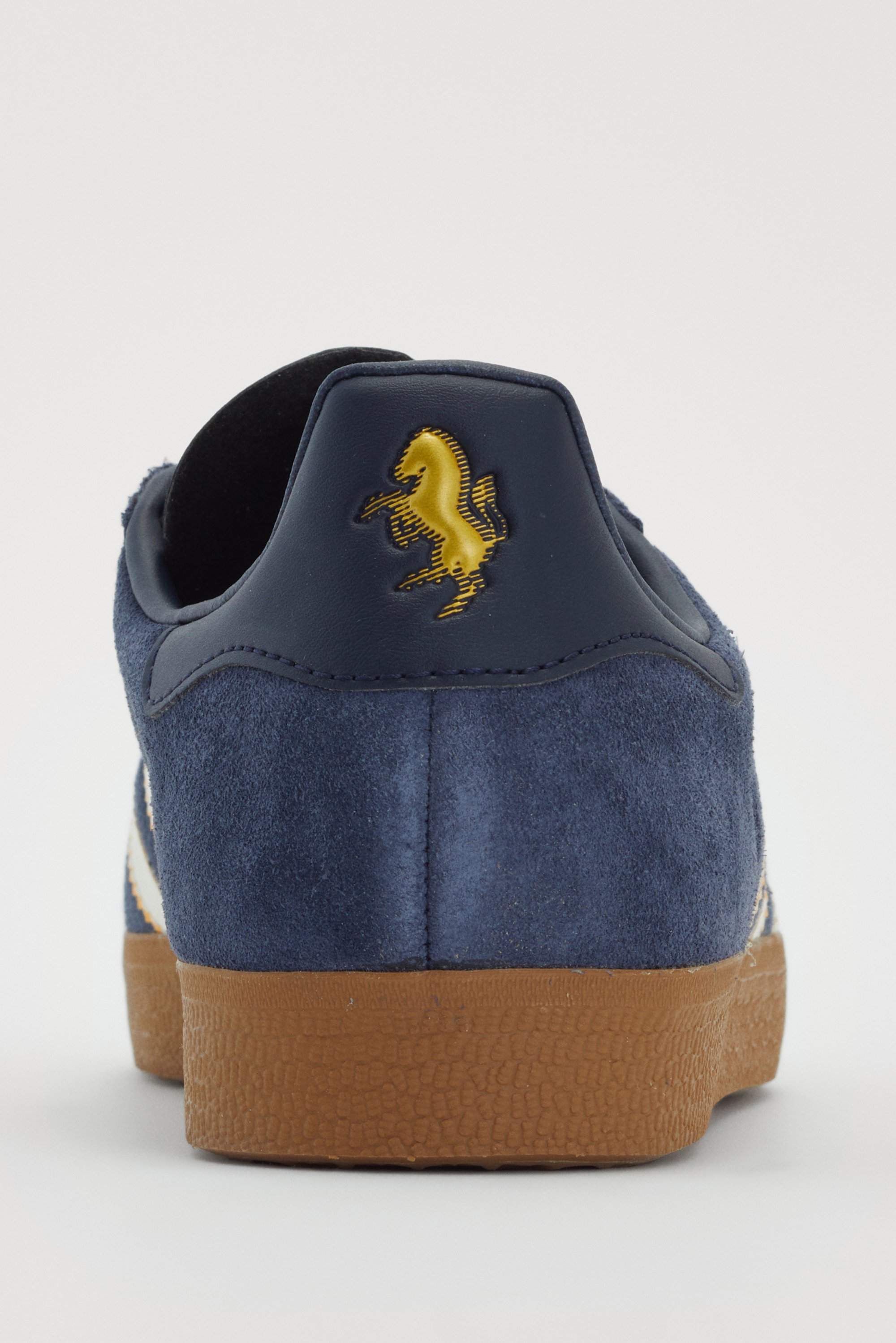 Navy suède sneaker met een gouden paardenlogo op de hiel en een gestructureerde bruine rubberen zool.
