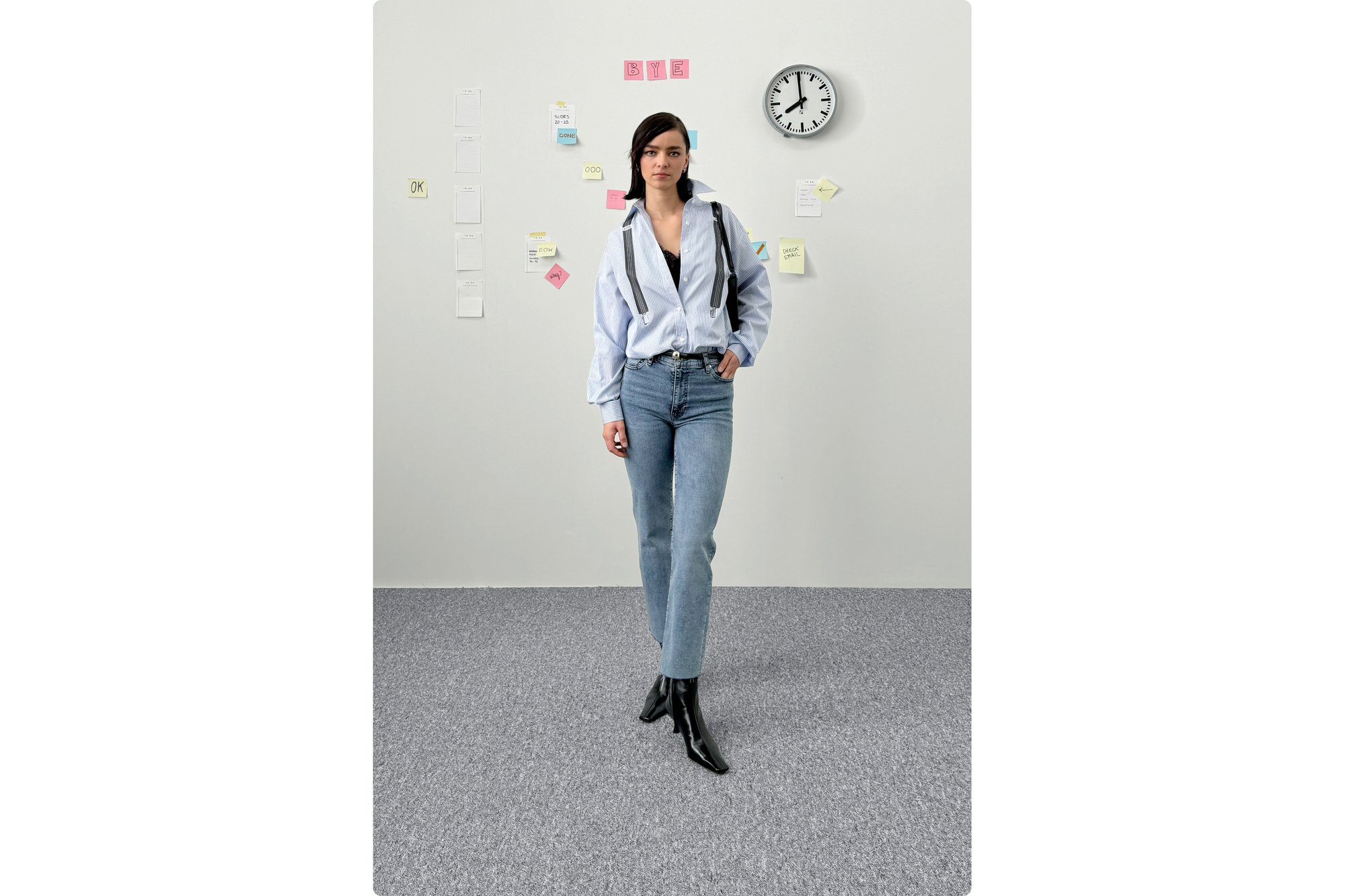 A woman in a striped blue shirt and black boots walks confidently in an office setting, with sticky notes and a clock on the wall.