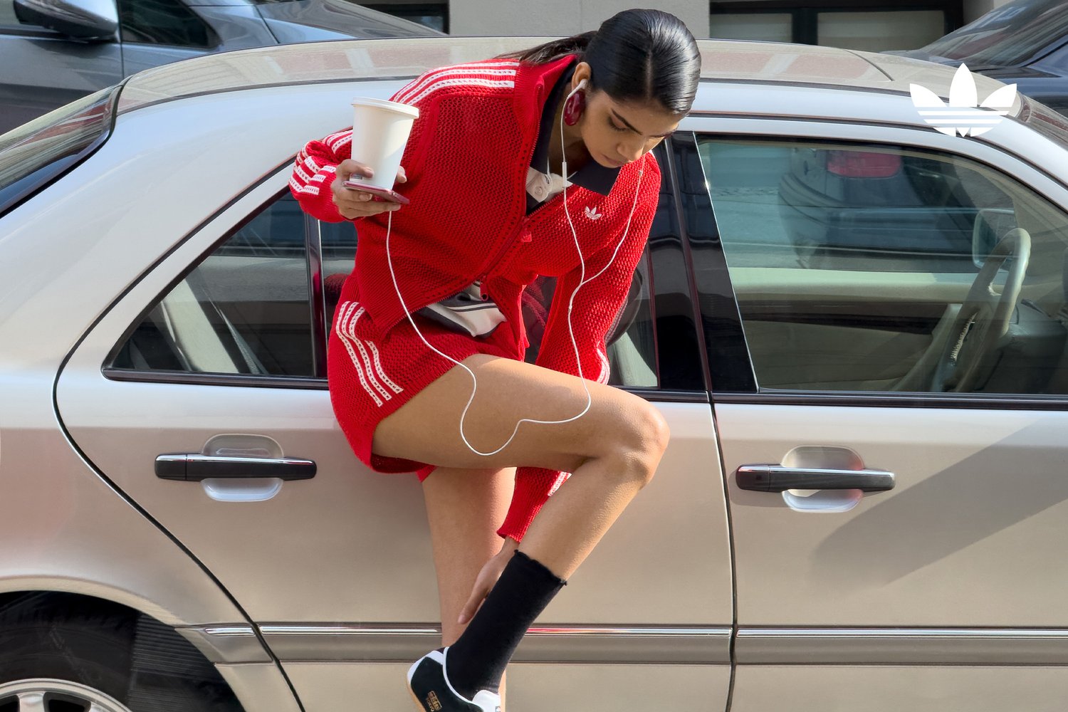 Een vrouw in een rode sportoutfit zit op de rand van een zilveren auto, houdt een koffiekop vast en draagt oordopjes terwijl ze gefocust naar haar telefoon kijkt.