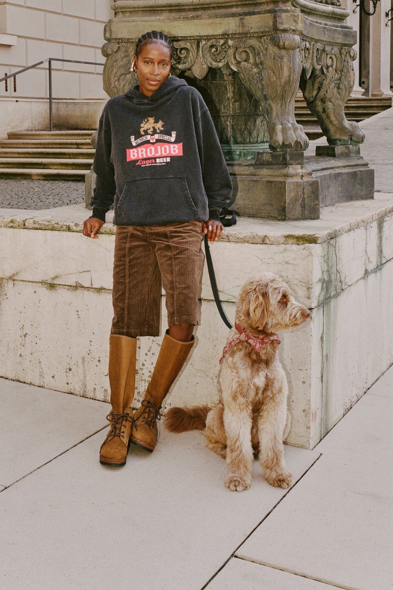 Zwarte hoodie met logo, bruine corduroy shorts en kniehoge bruine laarzen; een lichtbruine doodle hond met een rode bandana zit ernaast.