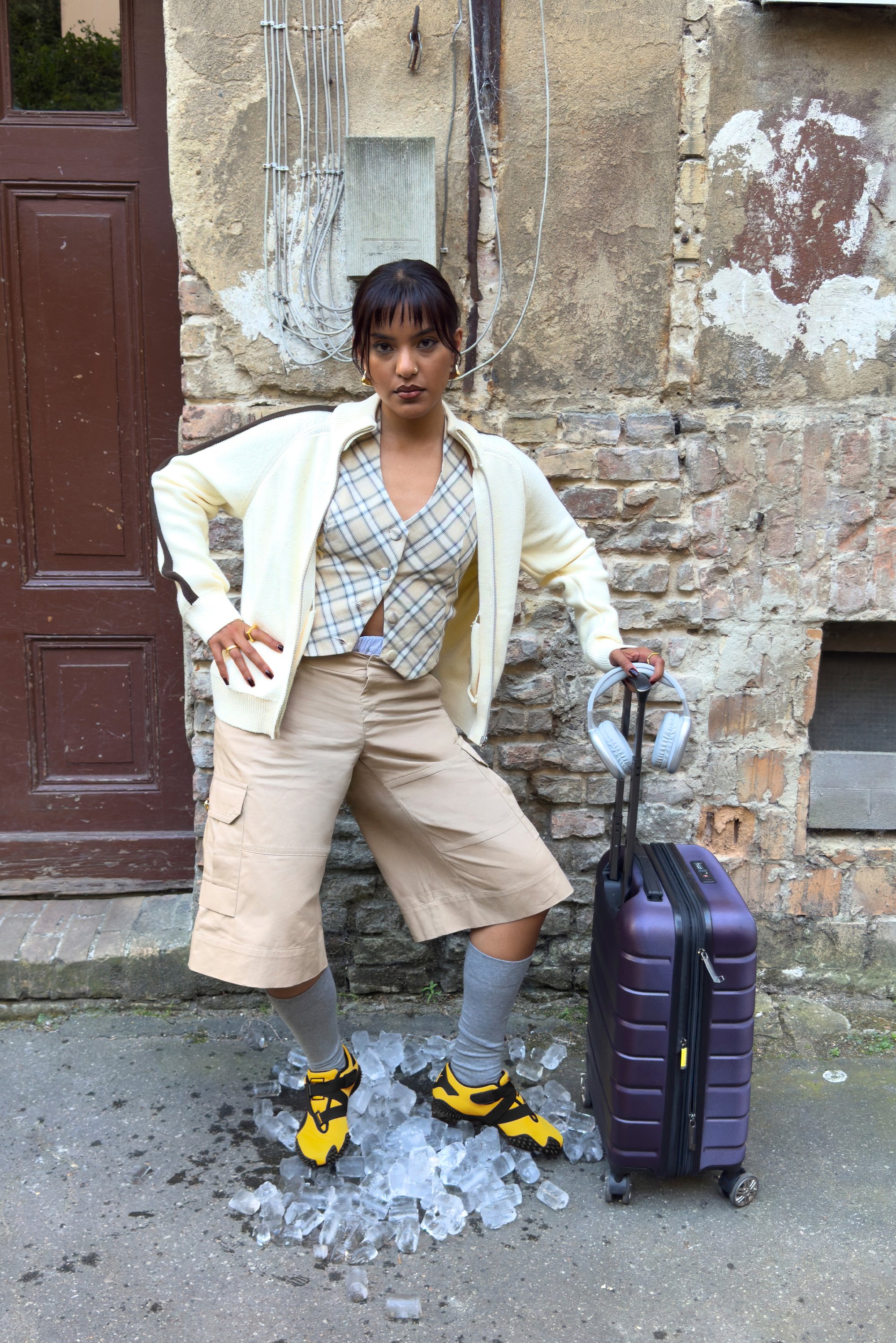 Maleta rígida morada junto a una persona que lleva una camisa de cuadros beige, zapatos amarillos y negros, y pantalones cortos de carga beige, de pie sobre cubitos de hielo.