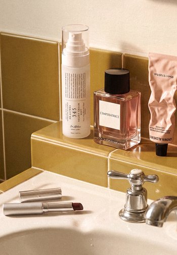 Évier de salle de bain avec robinet argenté, un rouge à lèvres rouge foncé, un spray pour le soin de la peau, un flacon de parfum rose et un tube de crème pour les mains froissé sur une crédence carrelée jaune moutarde.