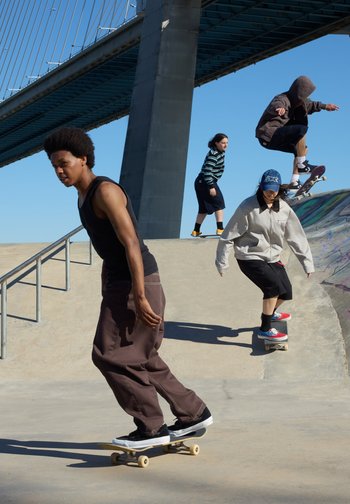 Four people skateboarding and riding under a large bridge on a sunny day at an urban skate park.