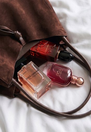 Sac à main en daim marron avec trois bouteilles de parfum partiellement à l'intérieur, posé sur une surface en tissu blanc.