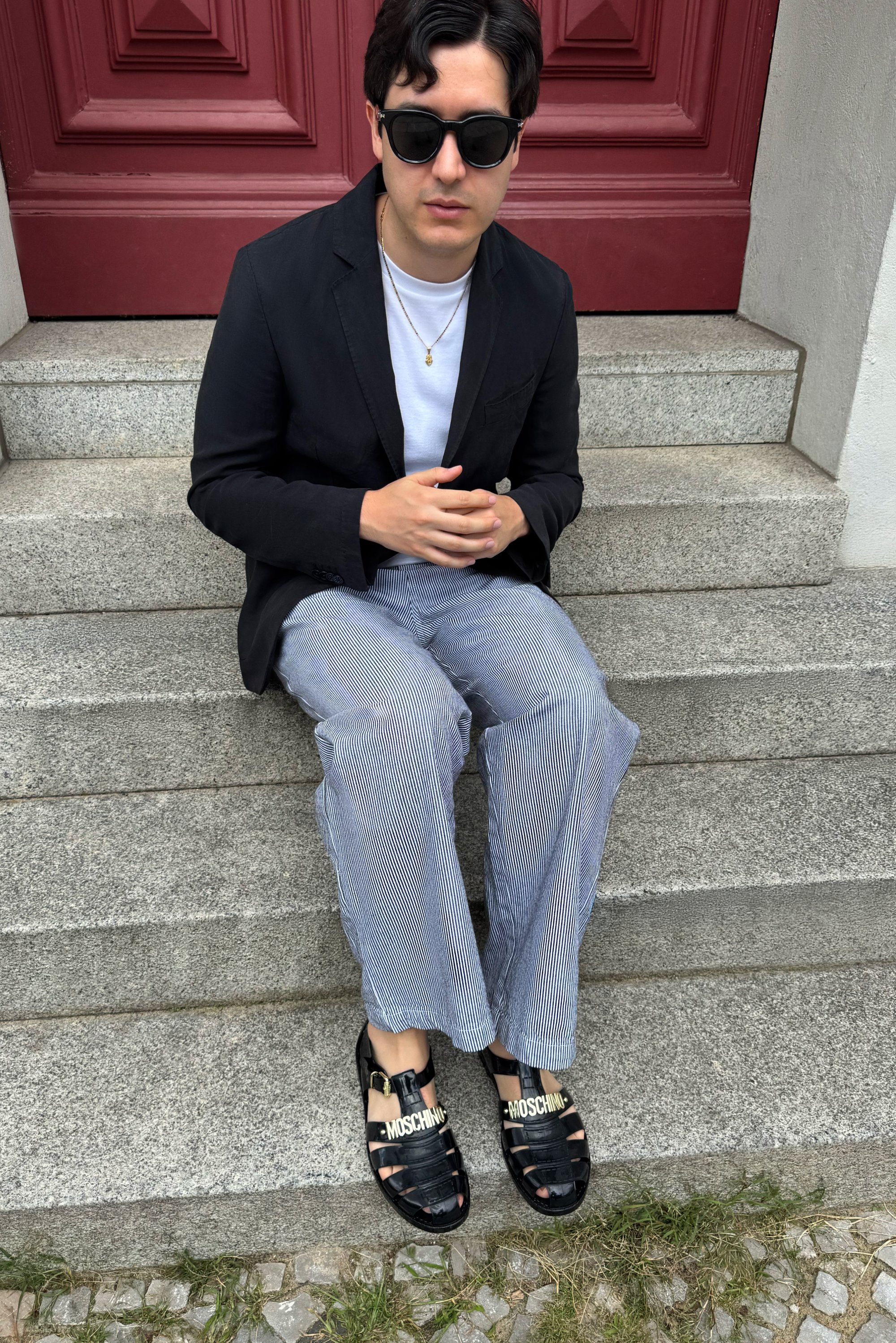Blazer nero su una camicia bianca, abbinato a pantaloni a righe. Indossa sandali neri con il marchio "MOSCHINO". Seduto su gradini di pietra.