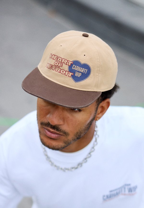 Beige cap med brun brem, med "HEART OF DETROIT" i rødt og "CARHARTT WIP" i blå hjertegrafikk; laget av bomull.
