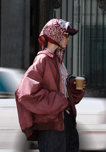 Overgrote bordeauxrode bomberjack, witte bikini top met strik, gestreepte rode hoofddoek, houdt een bruine koffiekop vast, spijkerbroek, stedelijke achtergrond.