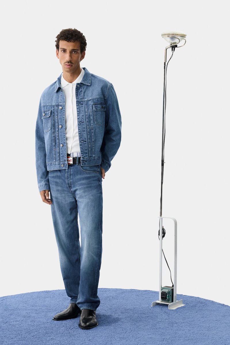 Denimjack en jeans in lichtblauw, met twee voorzakken. Model staat op een blauwe tapijt naast een vloerlamp met een witte kap en snoer.