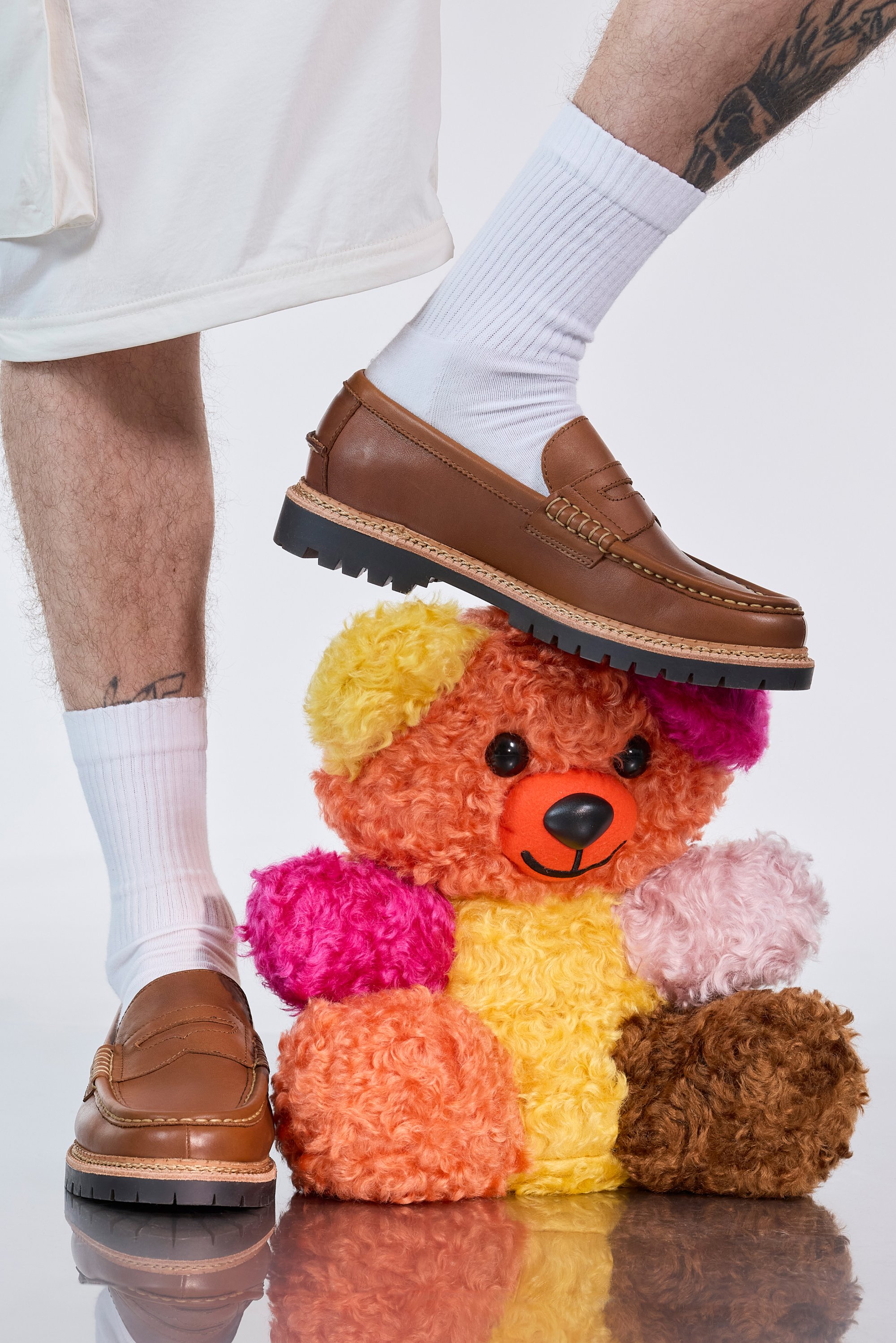 A person's foot in a brown shoe is resting on a colourful, fluffy teddy bear, displaying white socks against a light background.