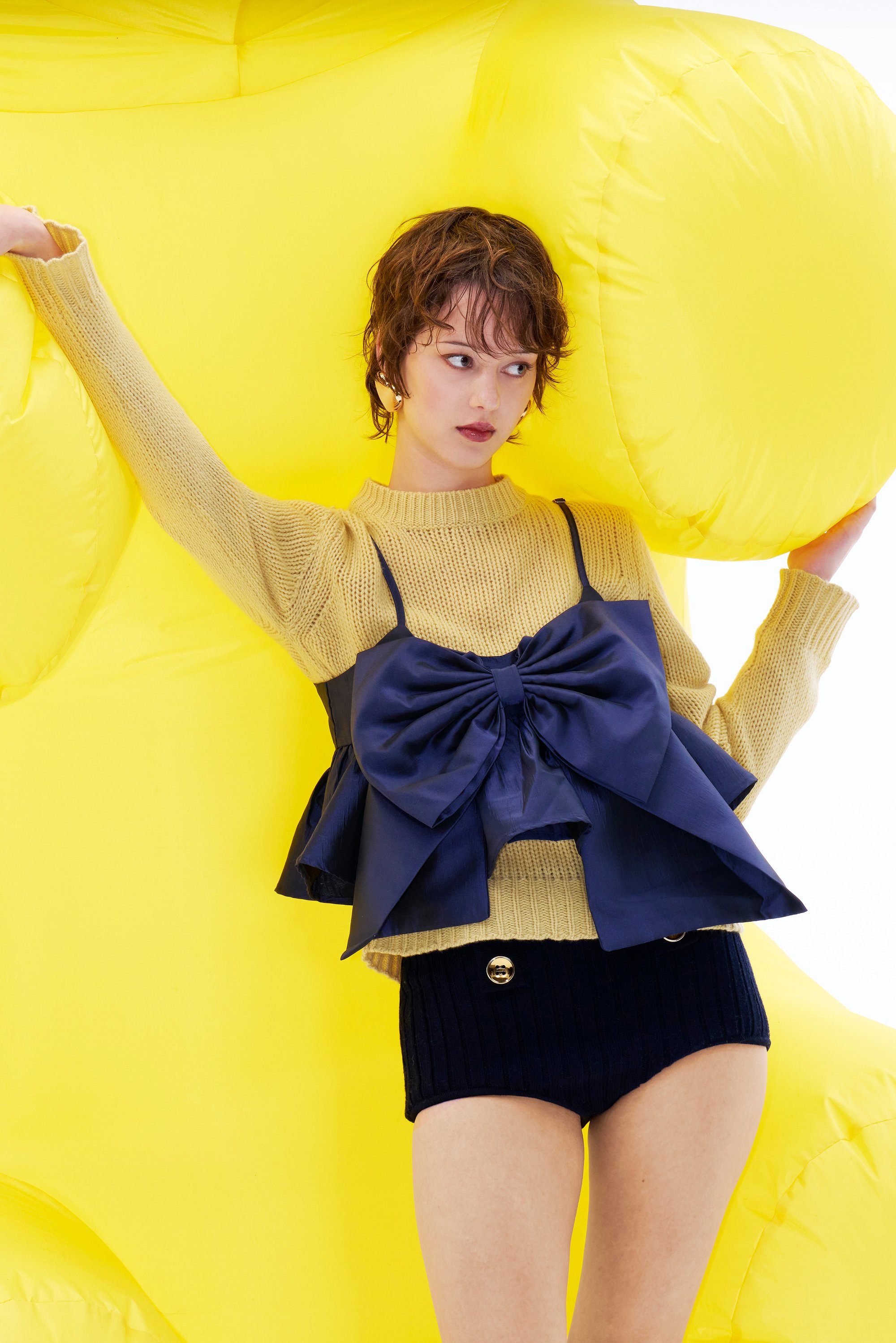 El modelo posa contra una gran estructura inflable amarilla, vistiendo un suéter amarillo de punto y una blusa azul marino con un lazo, combinada con pantalones cortos oscuros.