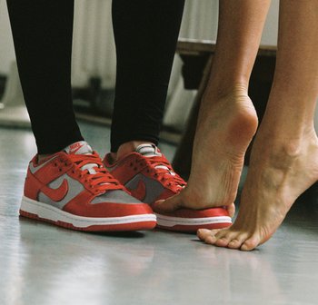 A foot in bare skin is positioned over a red and grey Nike sneaker, highlighting contrast between footwear and bare foot. Floor is light-colored.