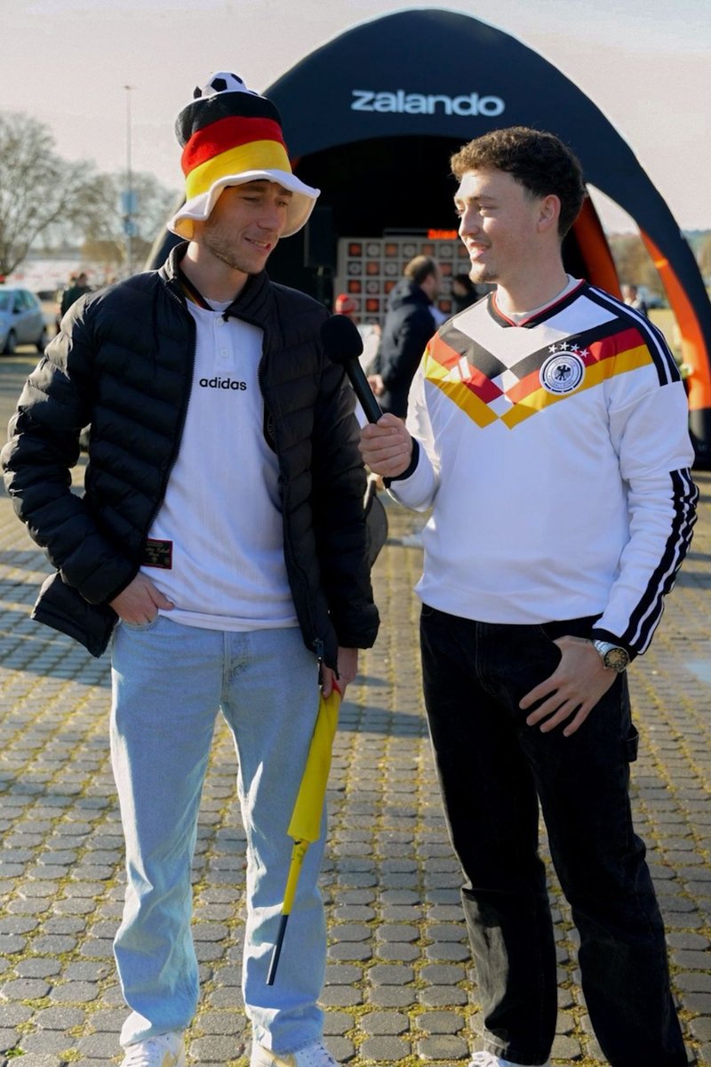 Two men in Germany-themed sportswear, one holding a microphone interviewing the other wearing a German flag hat near a Zalando booth outdoors.