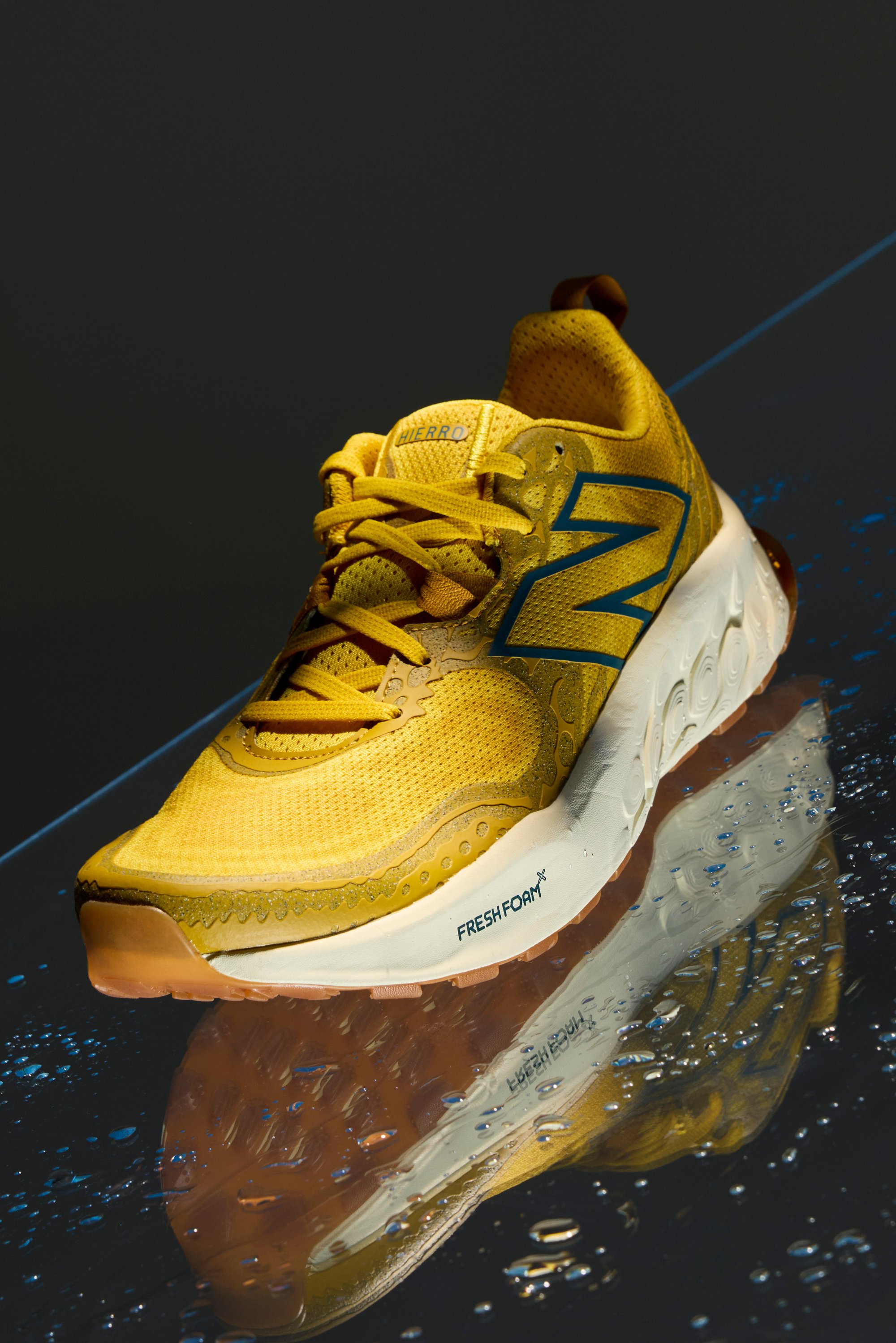 Zapatilla New Balance amarilla con parte superior de malla y detalles reflectantes, exhibida sobre un fondo oscuro con gotas de agua.