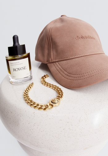 Eine weiche rosa Calvin Klein Mütze, eine Flasche mit organischem Hanföl mit Etikett und ein goldener Kettenarmreif werden auf einer gesprenkelten weißen Oberfläche präsentiert.