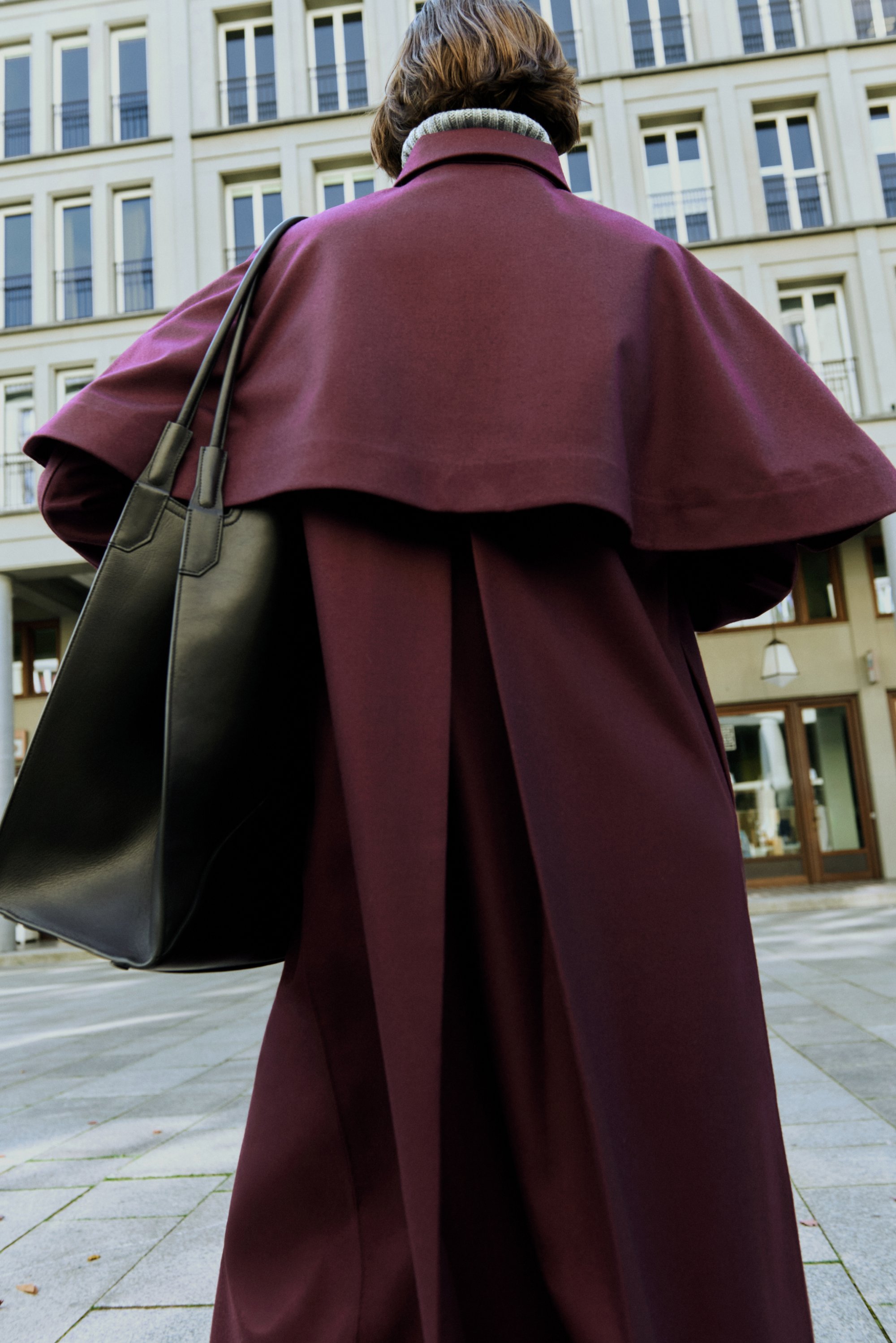 Langer burgunderfarbener Mantel mit einem geschichteten Cape-Design, kombiniert mit einer schwarzen Ledertasche. Glatte Textur; minimalistische Details.