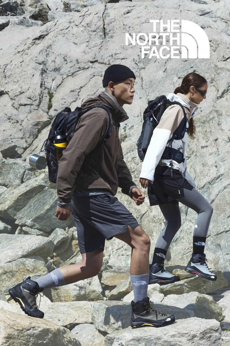 Two individuals traverse a rocky landscape. The man is wearing a brown jacket and shorts, while the woman is dressed in a colour-blocked jacket and grey leggings, both donning sturdy boots.