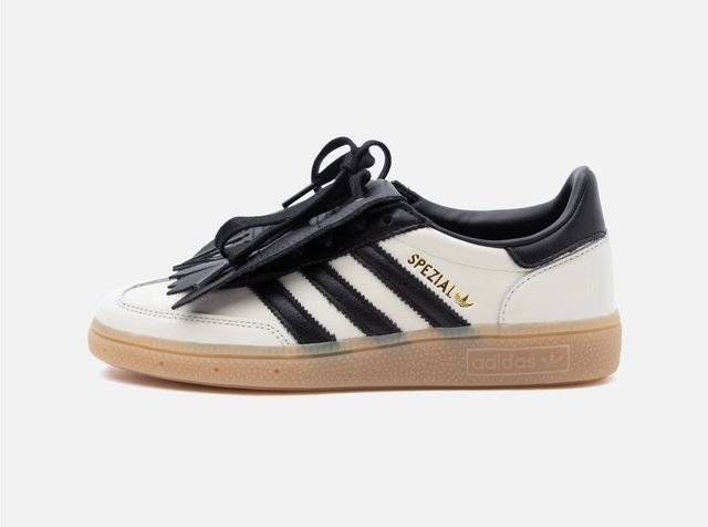 Zapatilla Adidas Spezial blanca y negra con suela de goma, cordones negros y flecos decorativos negros que cubren la lengüeta.